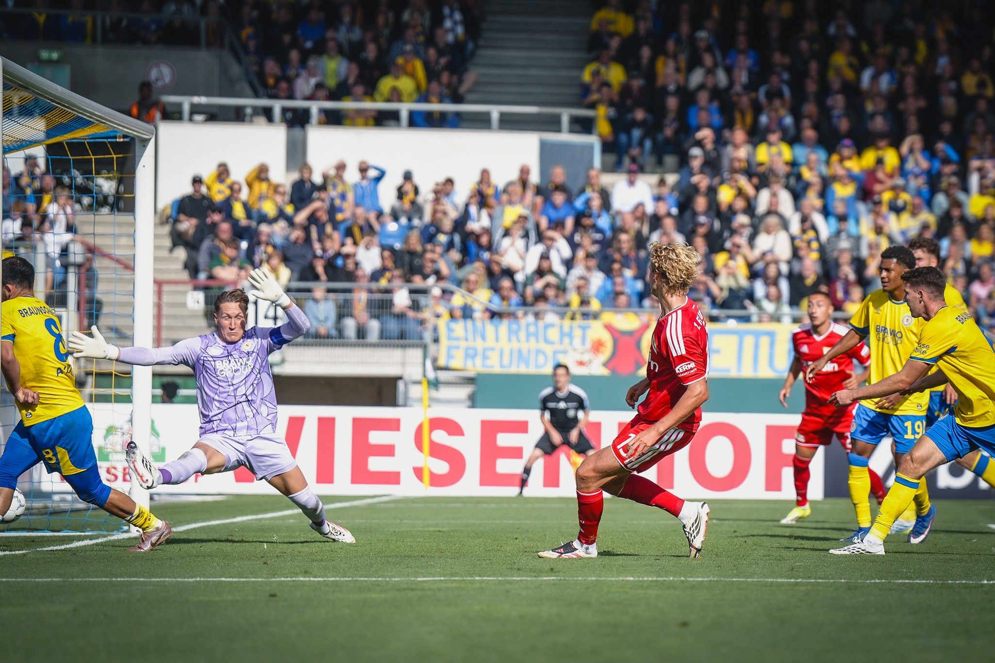 Ein Fußballspieler schießt auf das Tor, während der Torwart in einer Abwehrposition bereitsteht. Zuschauer im Hintergrund.