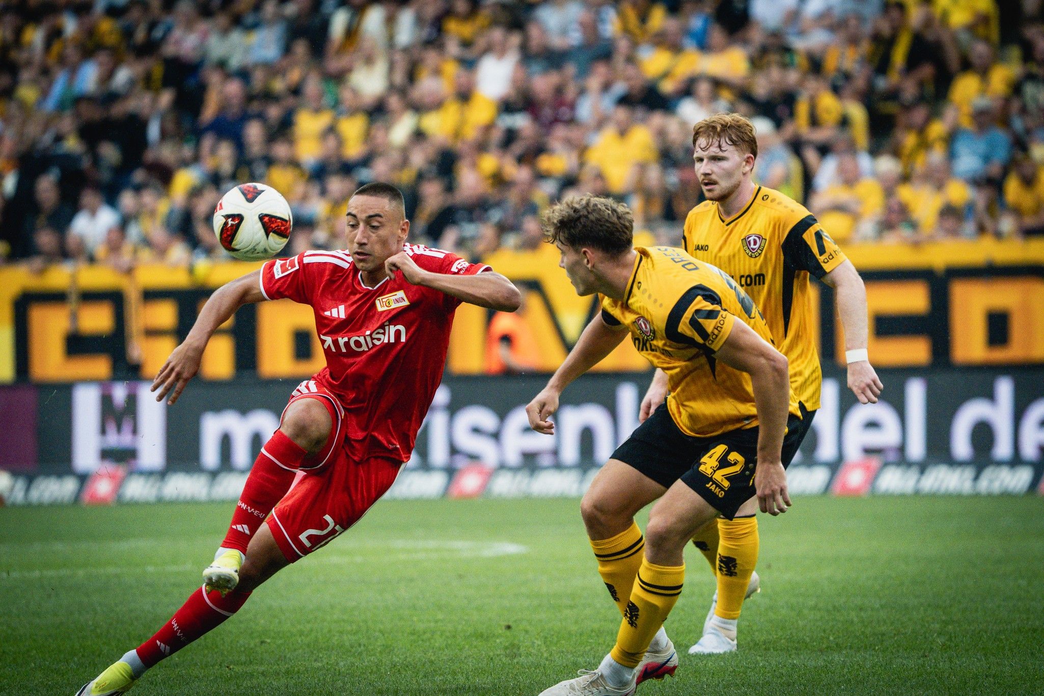 Ein Spieler in rotem Trikot tritt nach einem Ball, während zwei Gegner in gelben Trikots in der Nähe stehen.