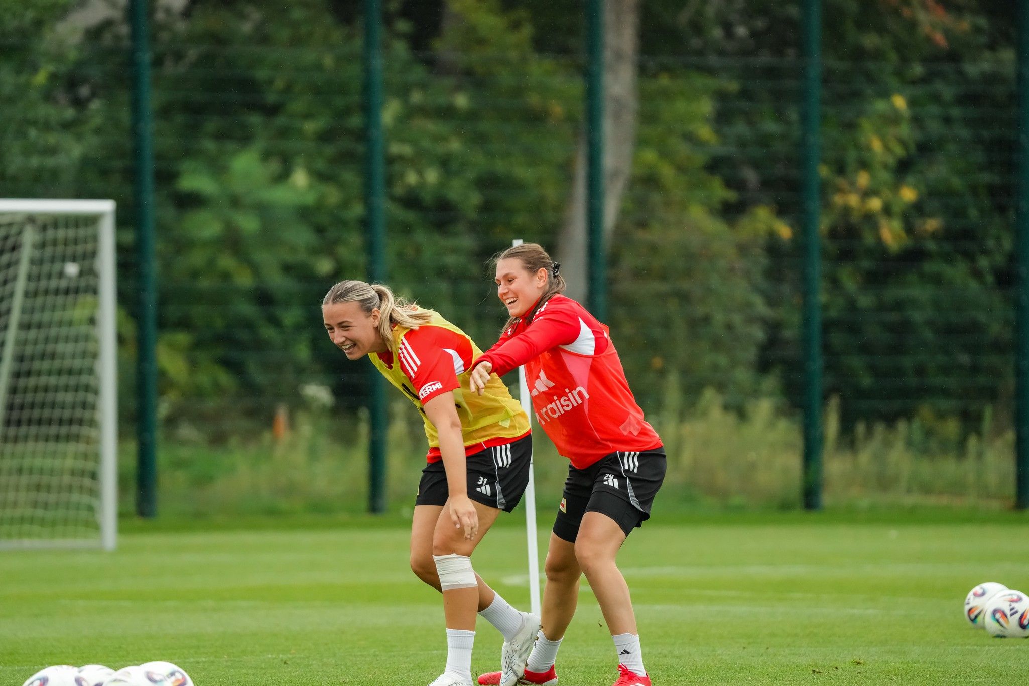 Zwei Fußballspielerinnen in Trainingstrikots lachen und interagieren auf dem Spielfeld, umgeben von Fußball.